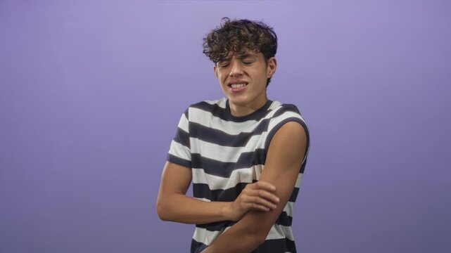 Teenage man clutching his left arm, rubbing his forearm with a pained expression while standing against a purple seamless studio backdrop; discomfort vulnerability.