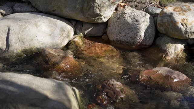 Clear water flows gently over smooth river stones. Sunlight glints softly on the moving surface. Rocks form a natural, rugged shoreline. Scene evokes calmness and quiet nature