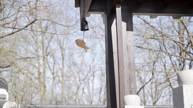 Wooden post holds small fish ornament. Fish dangles gently in breeze. Trees bare behind structure. Clear sky suggests early spring. Ideal for quiet, rustic scenes. Peaceful, uncluttered, natural feel