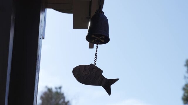Ancient bell hangs by chain against blue sky. Fish-shaped charm dangles below, catching breeze. Silent stillness suggests calm, peaceful moment. Ideal for serene, rustic, or spiritual themed projects