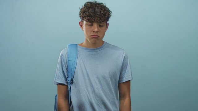 Teen boy with curly hair wearing light blue tee and backpack strap over shoulder looking sideways with puzzled expression in studio; adolescent uncertainty.