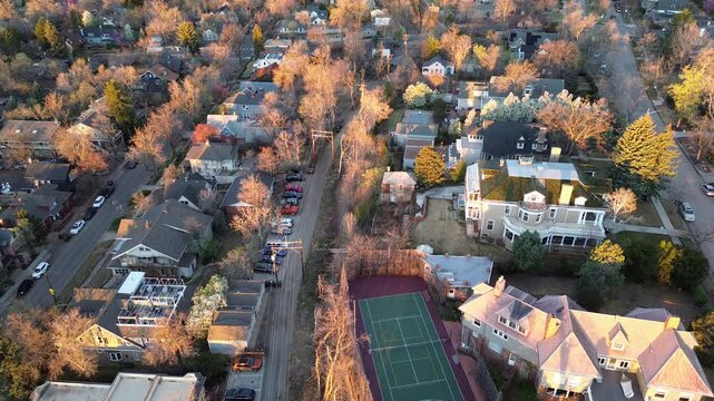 Central Boulder neighborhood shows tightly spaced homes, parked cars, fenced green sports court set among leafless street trees. Warm raking light over shingles, chimneys, driveways, pale trunks