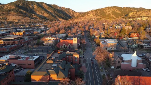 Historic downtown Boulder streets frame red roofed church tower, white roofed hall, compact blocks, Front Range slopes. Warm light glows over shingles, bare trees, mixed masonry by foothill ridges