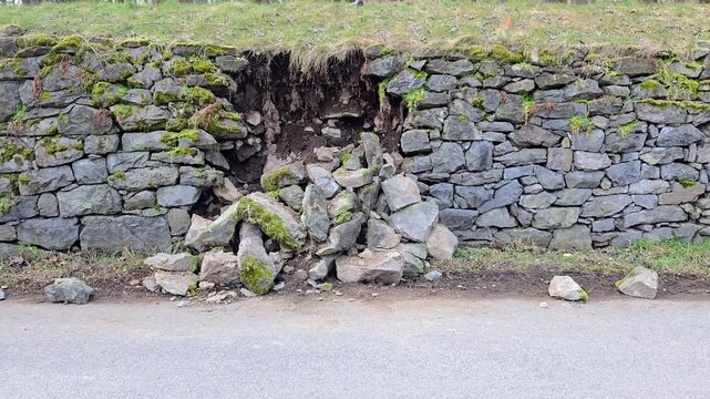 A detailed view of a partially collapsed dry stone retaining wall next to a rural asphalt road.
