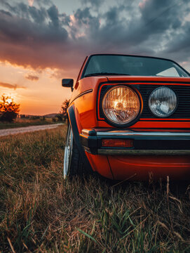 Vintage orange classic car parked on grassy field during a dramatic sunset with cloudy sky and rural countryside background scenery