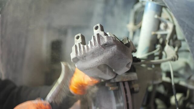 A close-up handheld video shows a mechanic wearing orange gloves cleaning brake components with a wire brush around the brake disc, removing rust and debris to ensure proper brake function, 2026