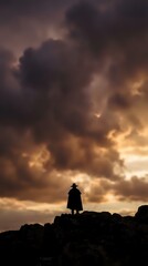 Fototapeta premium Silhouette of lone person standing on rocky cliff against dramatic stormy sky with golden sunset clouds for adventure and solitude concepts.