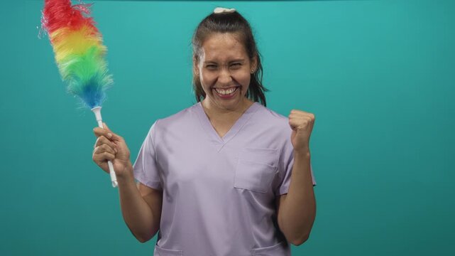 Woman cleaner holding rainbow duster and smiling with fist pump in teal studio setting for cleaning work; joy celebration.