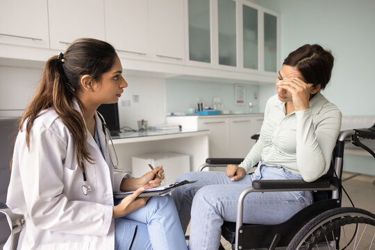 Young 30s Indian female psychologist or psychotherapist talks to worried woman in wheelchair during medical consultation, patient looks upset, need for psychological support, receiving disability care