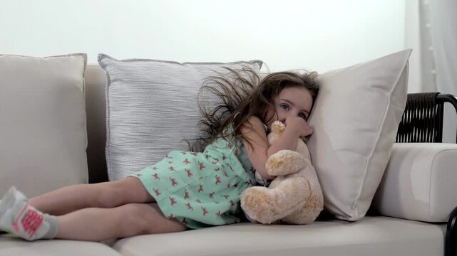 Cute young girl in a bright dress is lying on a big bright sofa and falling asleep hugging her stuffed toy