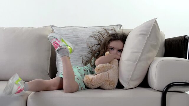 Young pretty little girl is lying on the couch with a stuffed toy and dangling her leg.