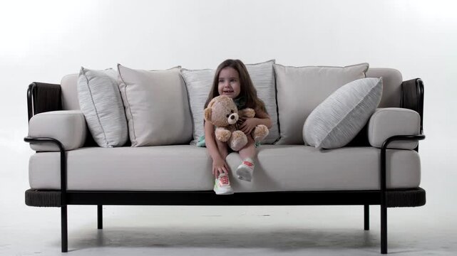 Beautiful little girl is sitting on a big bright couch with a soft toy in her hands and smiling. White background 