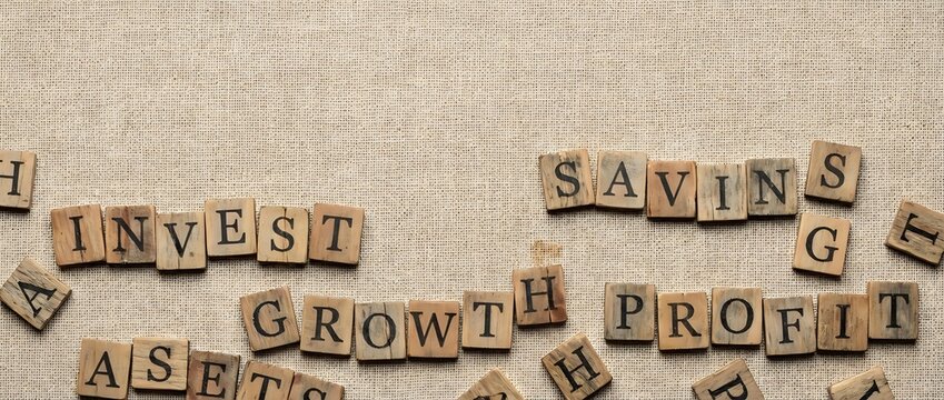 Wooden letter tiles spelling financial terms like invest, savings, growth, profit and assets scattered on textured beige background for business concepts.