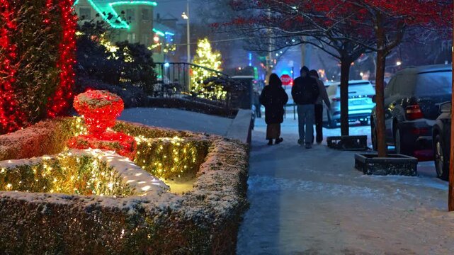People walking on snowy sidewalk past residential Christmas light display. Night view of snowy New York street with glowing festive lights on hedges and people walking in distance.