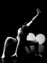 Fototapeta premium Woman sitting on the floor in dramatic dance pose with one arm raised, black and white, dark stage, two round lights in background. Expressive modern performance.