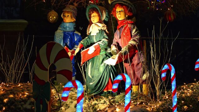 Three vintage Christmas caroler statues with glowing candy canes. Outdoor holiday display of singing figures in winter attire with snow at night in New York City.