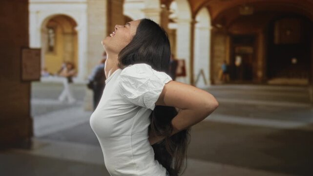 Woman holding lower back in sunlit historic old town stone building; backache discomfort strain tension.