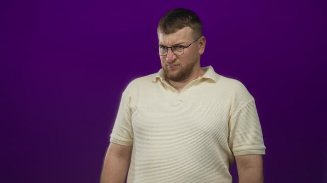 Man with glasses grimacing and squinting in studio against purple backdrop wearing cream polo shirt, hands down by sides; disgust discomfort.
