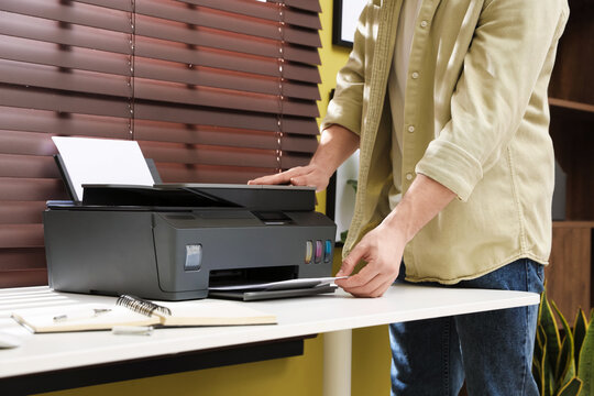 Man taking printed sheet of paper from printer at white desk indoors, closeup
