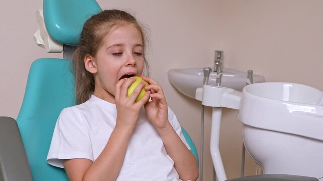 Little girl with long hair sits in a dental chair, taking a bite of a green apple while surrounded by dental equipment and a white sink in a modern clinic. Healthy teeth concept.