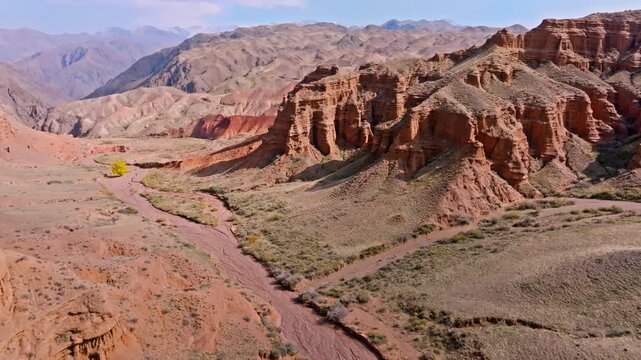 Adult man strolls through arid desert landscape with rocky canyons. Serene atmosphere enhanced by solitary yellow tree and dry riverbed.