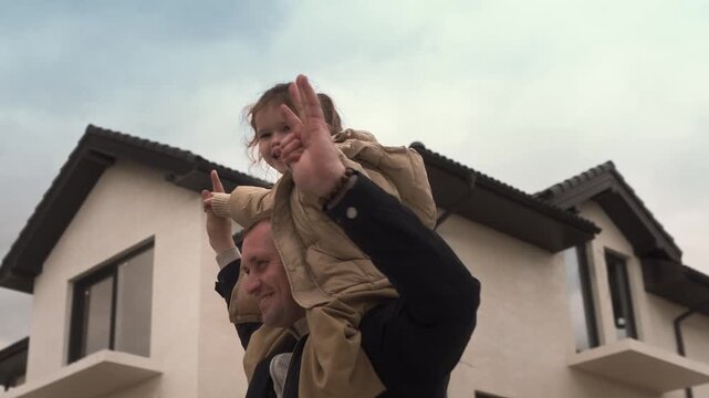 A successful middle-aged man rides his young daughter on his shoulders outside a large country house 