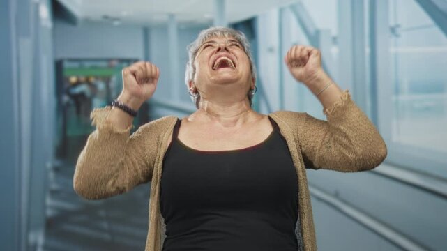 Woman raises fists in celebration on airport moving walkway while smiling and walking toward a gate; joy travel triumph.