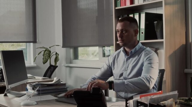 Manager with a laptop arrives at his workstation in the office, sits in his chair and looks at his watch 