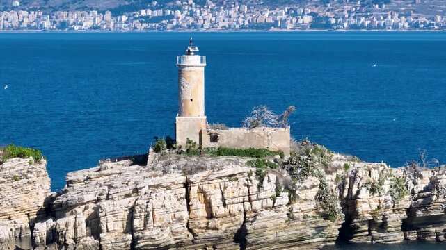 Aerial drone footage approaching the historic lighthouse on a rugged, rocky island. A cinematic view of a remote maritime beacon against the deep blue sea and distant city of Saranda