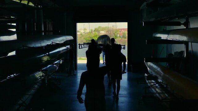 Silhouetted rowing crew carrying a shell outside the boathouse to the river for training