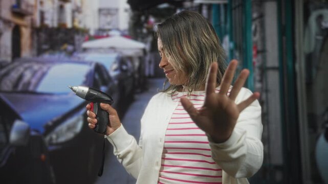 Hispanic middleaged woman holding cordless screwdriver, palm raised to stop, wincing face on a narrow street; unease caution.