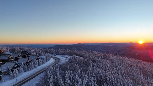 Aerial Drone View of Snowy Mountain Road and Ski Village at Sunset Winter Landscape with Sun Horizon

