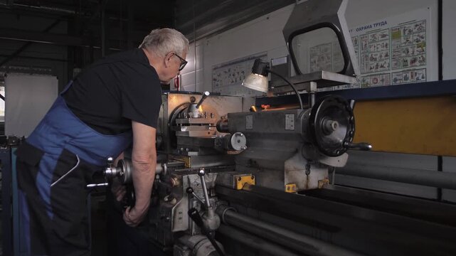 Elderly man in a work uniform works on a huge lathe