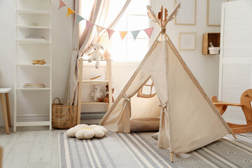 Stylish toy wigwam, furniture, pillows and decor near window in playroom. Interior design © New Africa