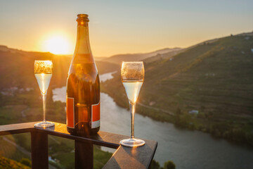 Naklejka premium Bottle of sparkling wine with two filled glasses overlooking the Douro River and vineyard hills at sunrise in the Douro Valley, Portugal.