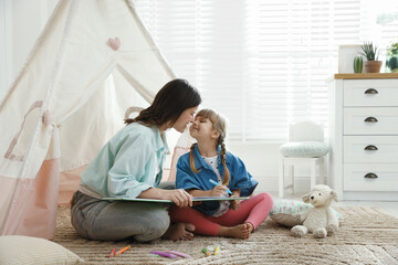 Mother and her daughter drawing in sketchbook together near toy wigwam at home, space for text © New Africa
