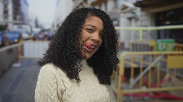 Woman wearing cream cable knit sweater sticks out tongue at a construction fence around a building under daylight; playfulness.