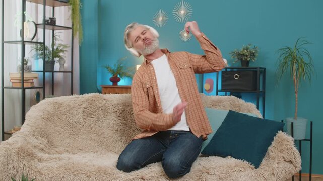 Happy Caucasian mature man kneeling on sofa at home, wearing wireless headphones. Smiling middle-aged guy energetically dances rock-n-roll, playing an imaginary guitar, enjoying the rhythm of music