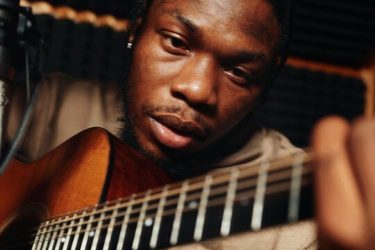 A musician in focused concentration plays acoustic guitar in a dark recording studio, a single spotlight illuminating visible sweat and string vibration in warm moody tones.