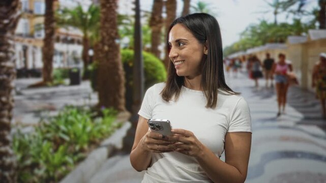 Woman holding smartphone with both hands on a palm lined street while smiling and glancing sideways; calm curiosity.