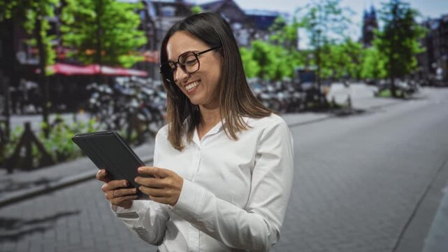 Young woman wearing white shirt and glasses holding tablet, hand touching screen and smiling on street lined with bicycles and trees; confidence joy.