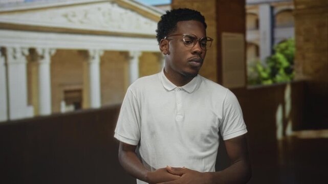 Man with clasped hands and contemplative gaze in building corridor with classical columns; introspective calm solitude reflection.