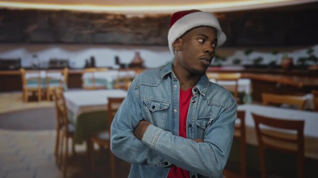 Man wearing santa hat crosses arms in building interior of casual restaurant with wooden chairs and tables; denial.