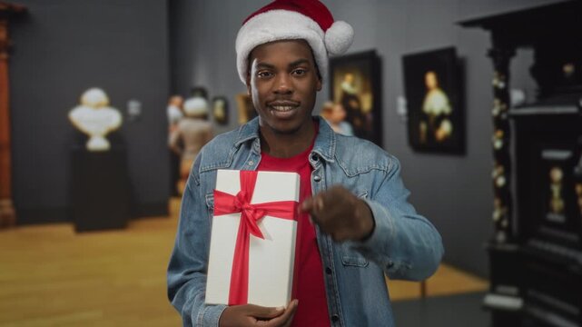 Man wearing santa hat and denim jacket holding white gift box wrapped in red ribbon and pointing finger in building; holiday joy.
