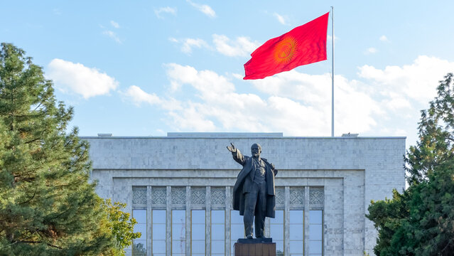 Lenin statue in Bishkek, Kyrgyzstan, with vibrant national flag waving against blue sky, showcasing historical significance and cultural identity