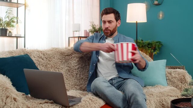 Adult man at home enjoys popcorn while watching movie on laptop, fully immersed in scene. Caucasian guy laughing in excitement at an unexpected moment, reacting emotionally to the thrilling storyline