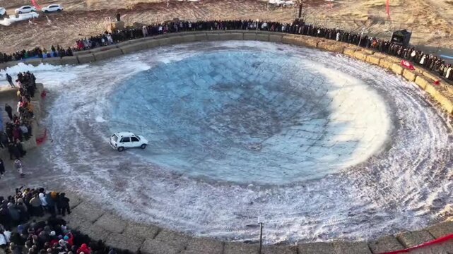 A wide horizontal shot capturing the immense scale of a man-made ice pit as a silver SUV circles the bottom, surrounded by hundreds of spectators in the Chinese countryside.
