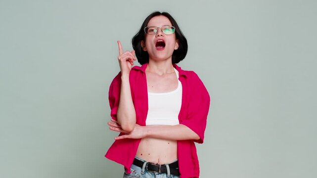 Young woman touches chin while thinking, then smiles and lifts finger up with excitement after finding idea or solution. Caucasian girl shows realization and inspiration, isolated on gray background.