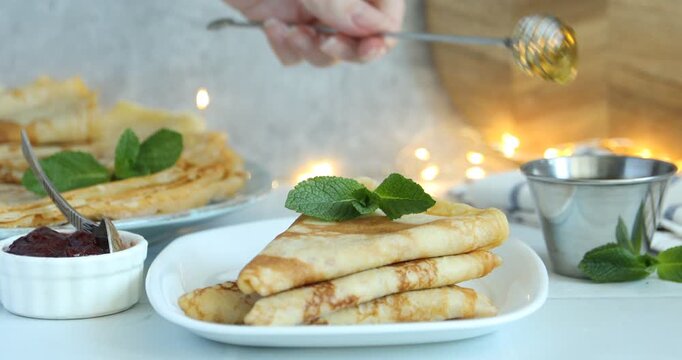 Woman pouring honey onto tasty crepes at white table, closeup
