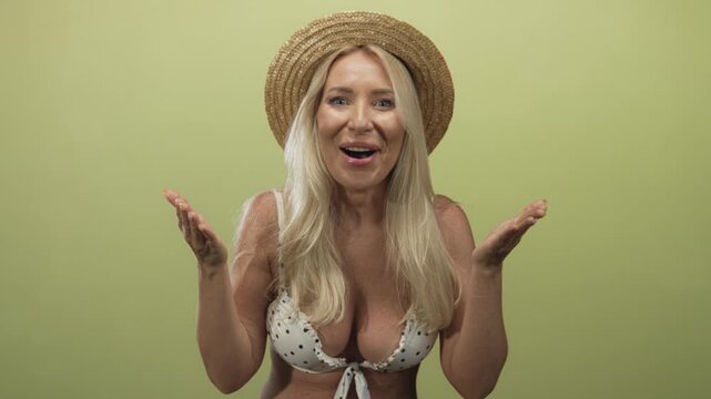 Woman raises open palms wearing straw hat and polka dot bikini top against green studio backdrop; surprise.